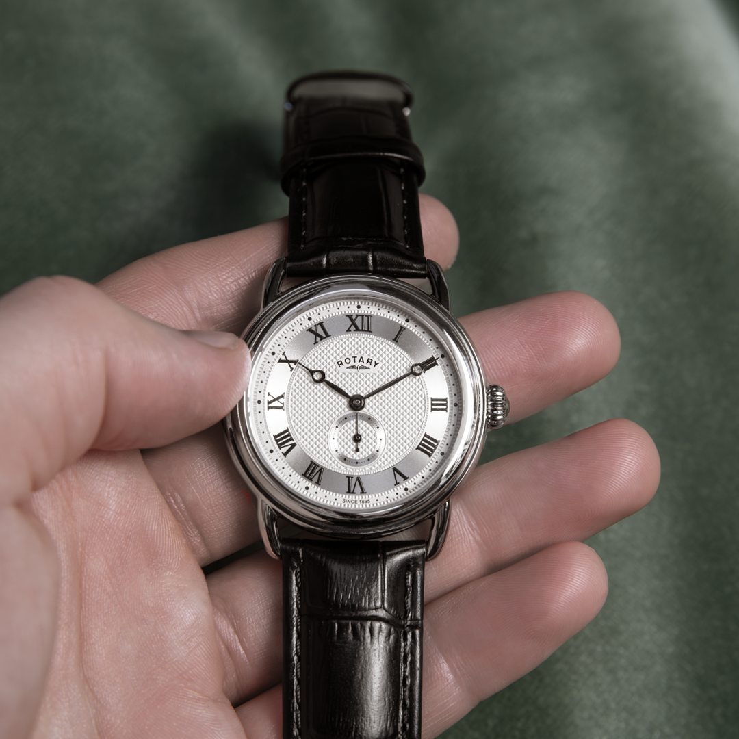 A man holding a Rotary sherlock watch with a stainless steel case, a silver dial with roman numerals and a black leather strap